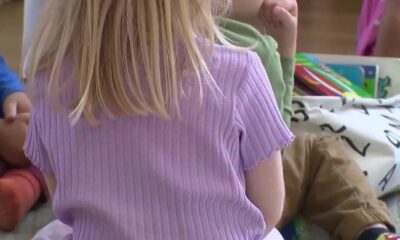 Back view of a child with long blonde hair in a purple ribbed shirt, sitting on the floor with books in a classroom setting.