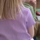 Back view of a child with long blonde hair in a purple ribbed shirt, sitting on the floor with books in a classroom setting.