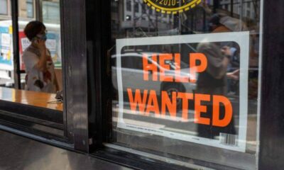 Shop storefront window with a bold 'HELP WANTED' sign; a masked person on the street is visible in the background reflection/through the glass
