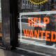 Shop storefront window with a bold 'HELP WANTED' sign; a masked person on the street is visible in the background reflection/through the glass