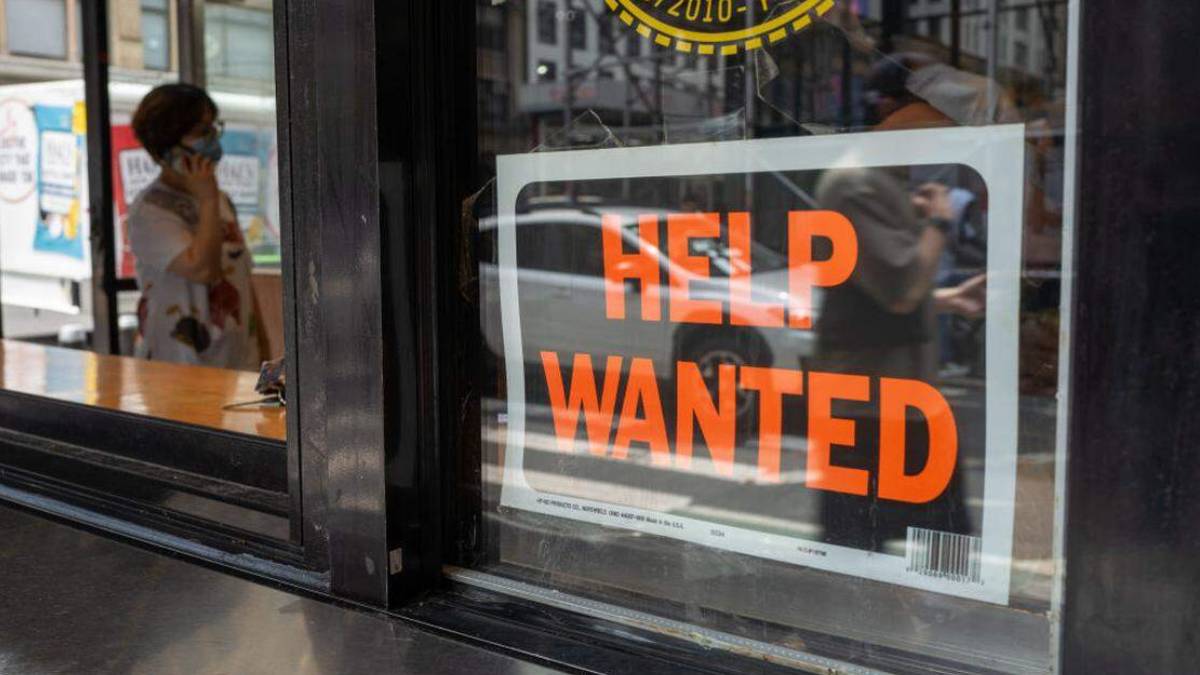 Shop storefront window with a bold 'HELP WANTED' sign; a masked person on the street is visible in the background reflection/through the glass
