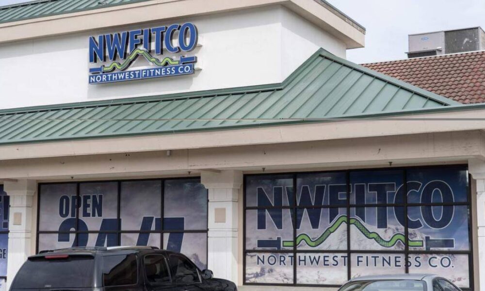 Front view of a fitness studio storefront with large NWFITCO/Northwest Fitness Co. sign on the façade and window graphics stating Open 24/7, plus parked cars in front.