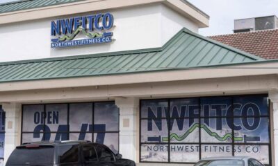 Front view of a fitness studio storefront with large NWFITCO/Northwest Fitness Co. sign on the façade and window graphics stating Open 24/7, plus parked cars in front.