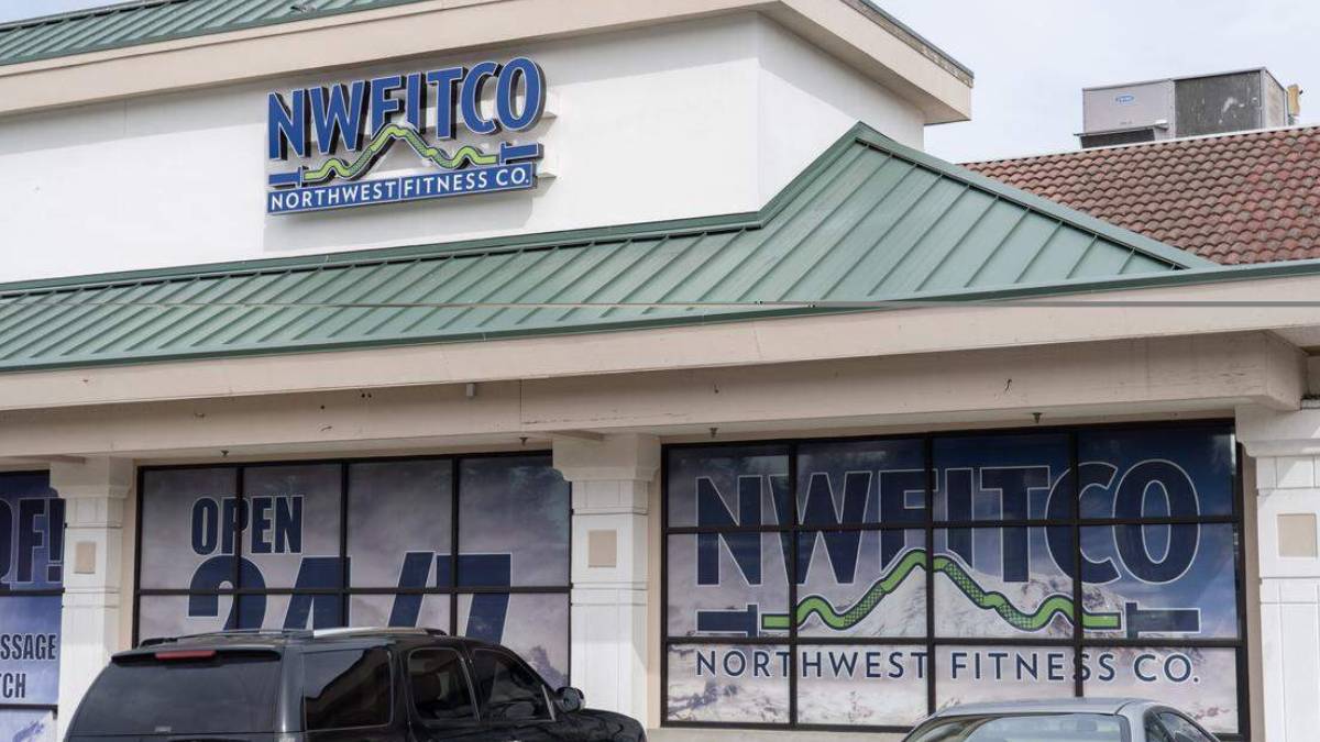 Front view of a fitness studio storefront with large NWFITCO/Northwest Fitness Co. sign on the façade and window graphics stating Open 24/7, plus parked cars in front.