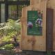 Wooden sculpture with a green poster about forests connecting us all; phrases include 'Our lungs' and 'Our happiness'. Outside a building entrance with a ramp visible to the left.