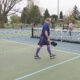 People playing pickleball on outdoor court, one man in purple reaching for ball near net while others watch in background