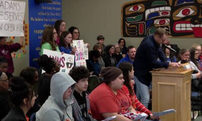Man in a dark sweater speaks at a wooden podium as a diverse audience watches a meeting, with students holding protest signs in the background.