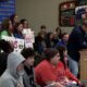 Man in a dark sweater speaks at a wooden podium as a diverse audience watches a meeting, with students holding protest signs in the background.