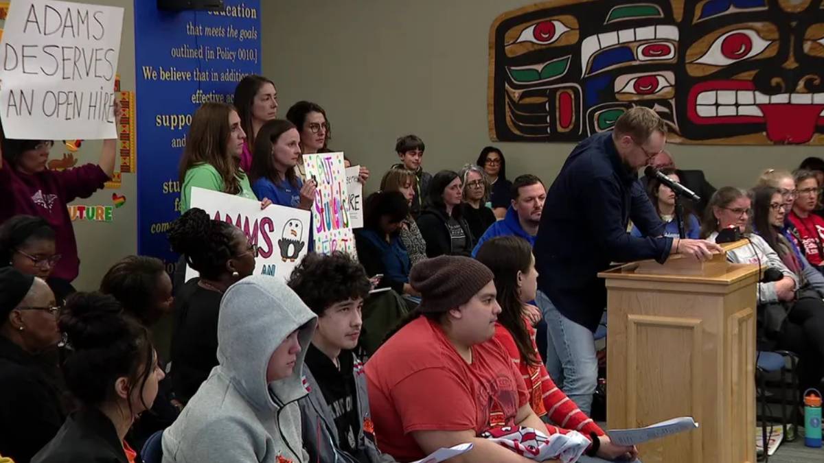 Man in a dark sweater speaks at a wooden podium as a diverse audience watches a meeting, with students holding protest signs in the background.