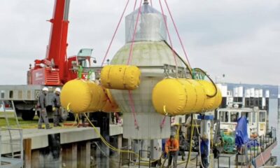Crane lowers a gray offshore module on a dock, surrounded by yellow inflatable pontoons and workers in hard hats.