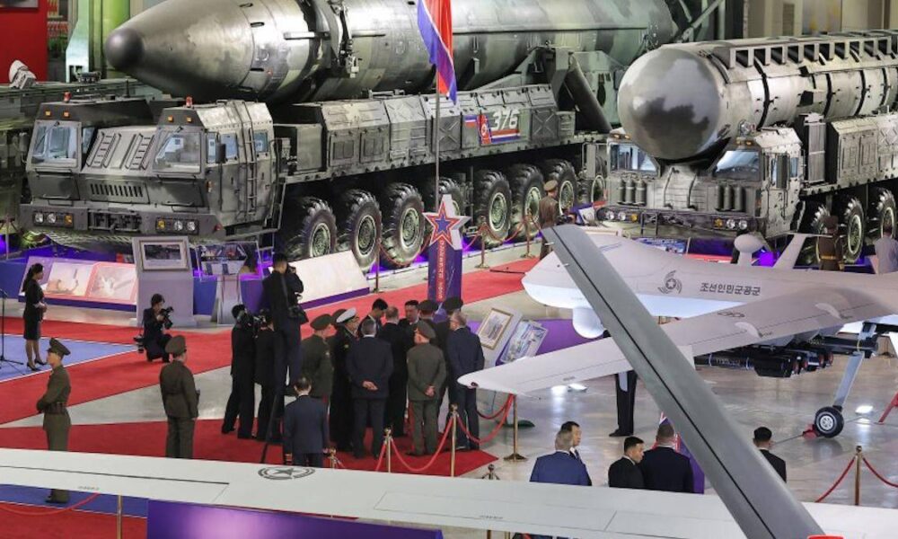 Military exhibit: large missile launcher trucks and a jet on display with officials and photographers in a hangar-like venue on a red carpet.