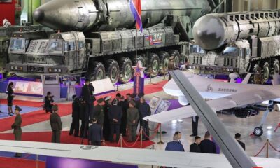 Military exhibit: large missile launcher trucks and a jet on display with officials and photographers in a hangar-like venue on a red carpet.