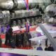 Military exhibit: large missile launcher trucks and a jet on display with officials and photographers in a hangar-like venue on a red carpet.