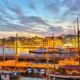 Harbor at dusk with moored sailboats, calm water reflecting warm city lights and a glowing skyline.