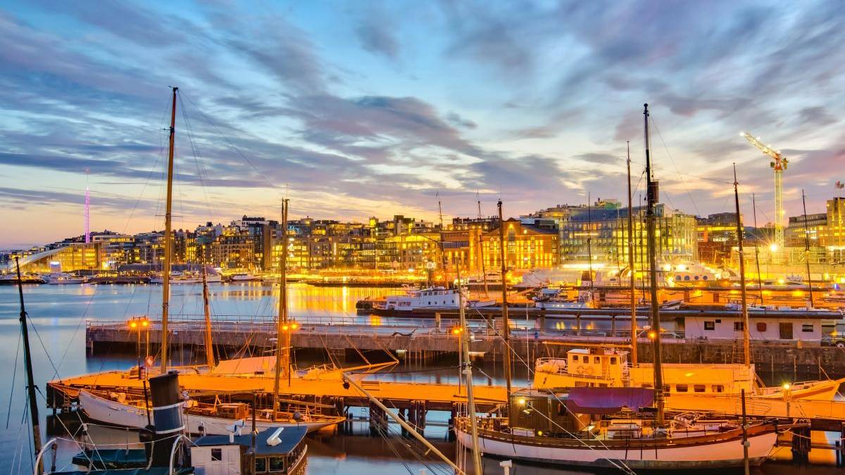 Harbor at dusk with moored sailboats, calm water reflecting warm city lights and a glowing skyline.