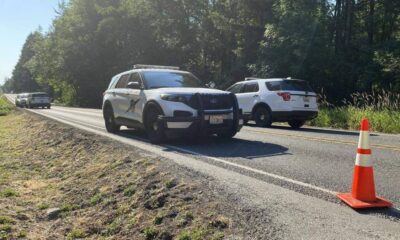 Two police SUVs are stopped on a rural highway with an orange traffic cone in the foreground and a forested shoulder nearby.