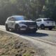 Two police SUVs are stopped on a rural highway with an orange traffic cone in the foreground and a forested shoulder nearby.
