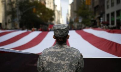 Soldier in camouflage uniform seen from behind, standing before a large waving American flag in a city street parade.