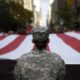 Soldier in camouflage uniform seen from behind, standing before a large waving American flag in a city street parade.