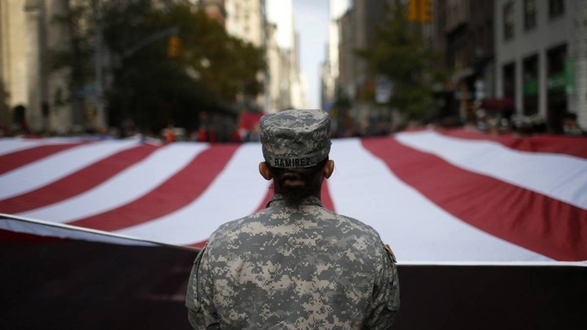 Soldier in camouflage uniform seen from behind, standing before a large waving American flag in a city street parade.