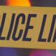 Close-up of yellow police line tape with bold black text POLICE LINE, likely at a crime scene beneath colored lights in the background.
