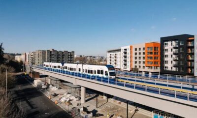 Modern white light-rail train on an elevated track with colorful apartment blocks in the background under a clear blue sky.