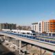 Modern white light-rail train on an elevated track with colorful apartment blocks in the background under a clear blue sky.