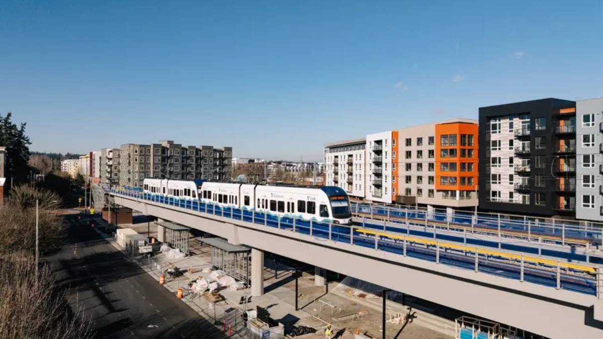 Modern white light-rail train on an elevated track with colorful apartment blocks in the background under a clear blue sky.