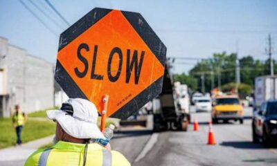 Worker in high-visibility clothing holds an orange 'SLOW' sign at a roadside construction zone, with traffic cones and vehicles in the background.