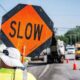 Worker in high-visibility clothing holds an orange 'SLOW' sign at a roadside construction zone, with traffic cones and vehicles in the background.