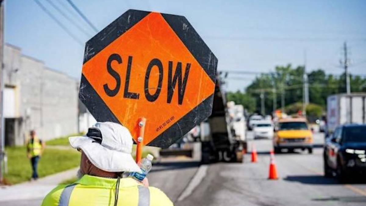 Worker in high-visibility clothing holds an orange 'SLOW' sign at a roadside construction zone, with traffic cones and vehicles in the background.