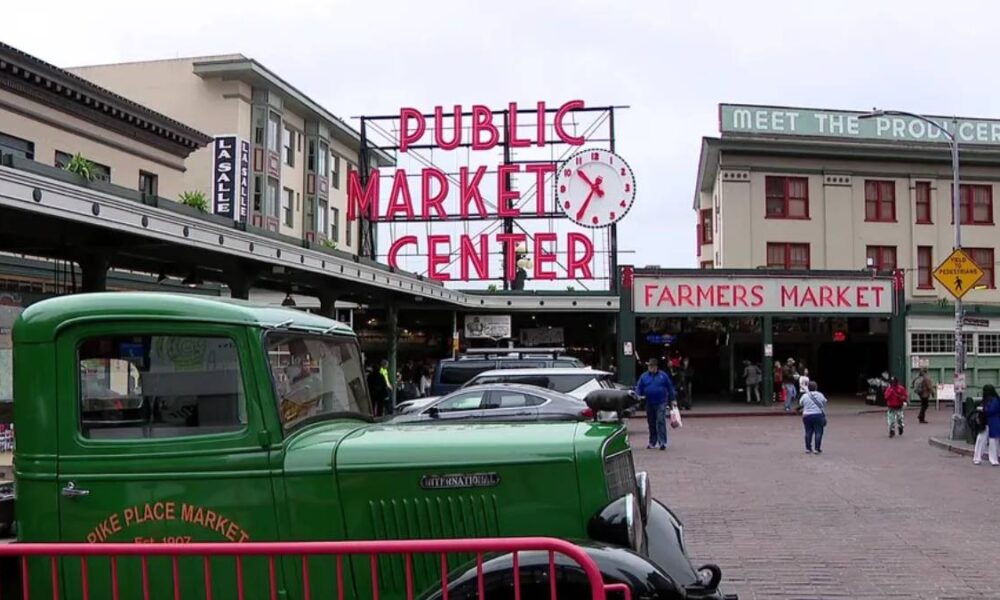 Green vintage truck parked in front of the Public Market Center entrance, with a Farmers Market sign and people walking nearby.