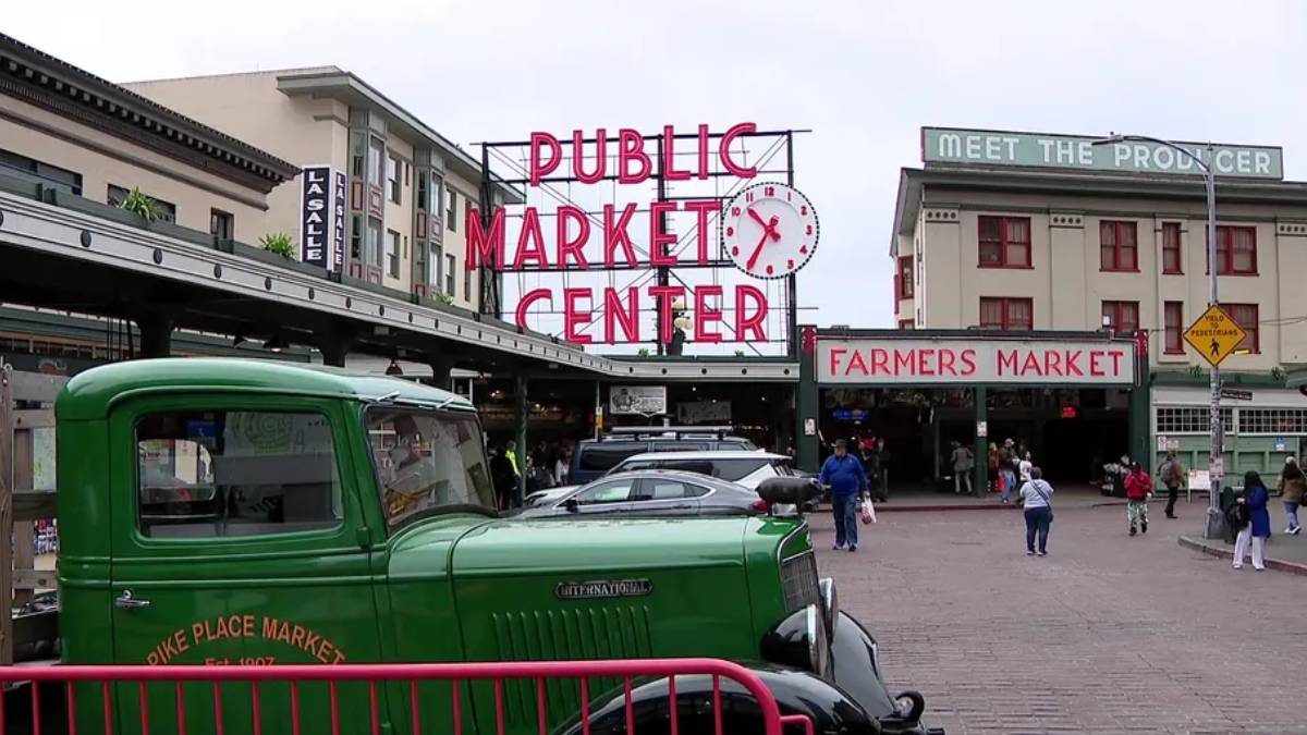 Green vintage truck parked in front of the Public Market Center entrance, with a Farmers Market sign and people walking nearby.