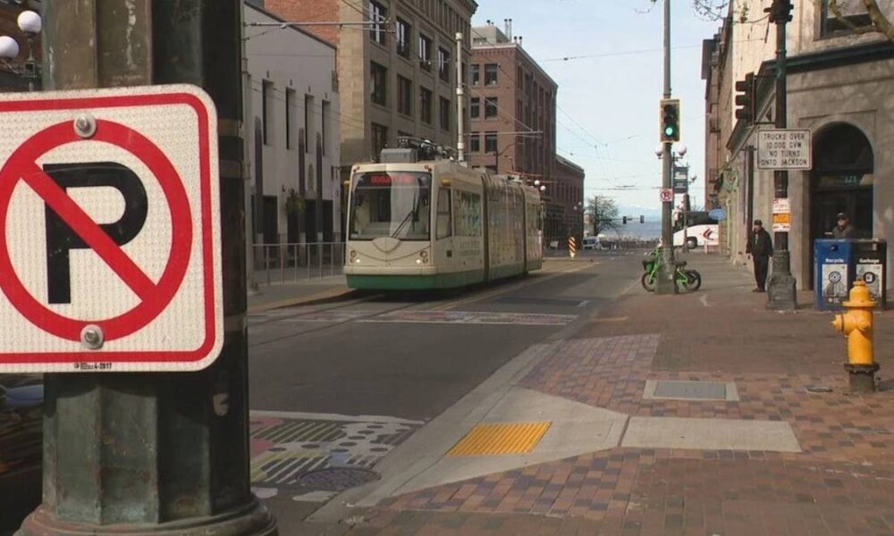 Tram on a city street seen from the sidewalk with a large no-left-turn sign in the foreground and pedestrians nearby.