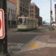 Tram on a city street seen from the sidewalk with a large no-left-turn sign in the foreground and pedestrians nearby.