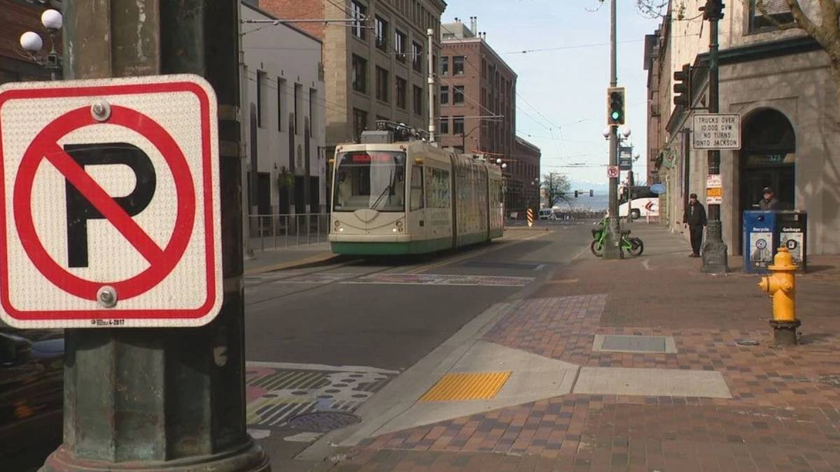 Tram on a city street seen from the sidewalk with a large no-left-turn sign in the foreground and pedestrians nearby.