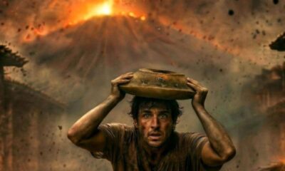 Man wearing a muddy shirt holds a clay pot on his head while chaos and lava glow behind him during an eruption.