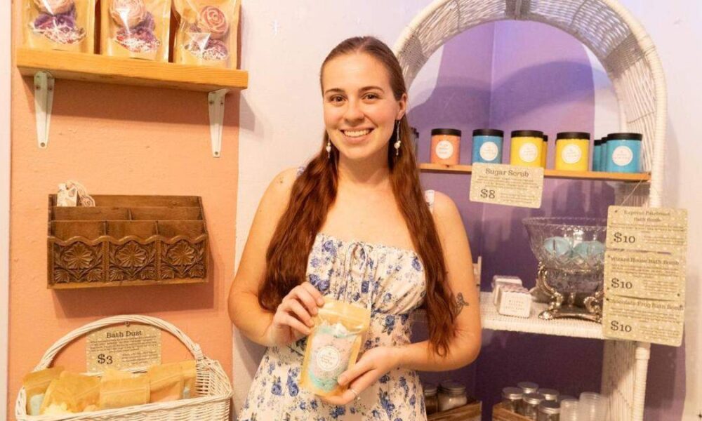 Woman in a pastel shop holding a bag of bath product, smiling at the camera with shelves of soaps behind her.