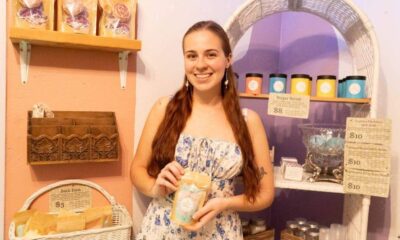 Woman in a pastel shop holding a bag of bath product, smiling at the camera with shelves of soaps behind her.