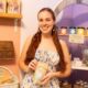 Woman in a pastel shop holding a bag of bath product, smiling at the camera with shelves of soaps behind her.