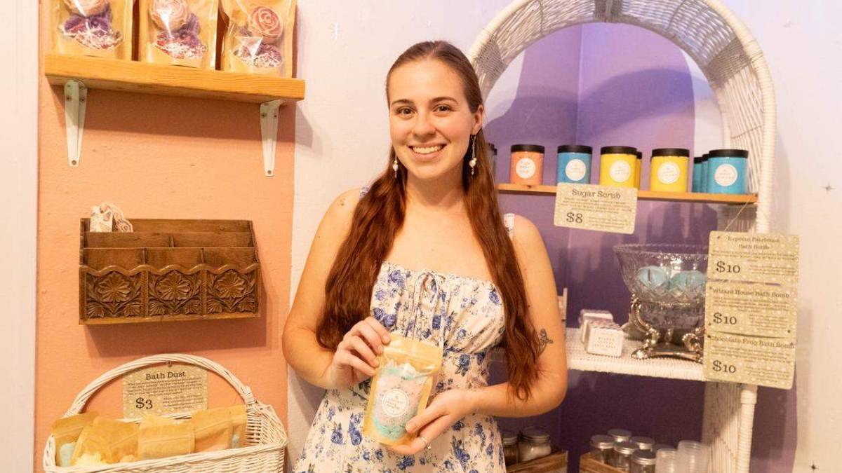 Woman in a pastel shop holding a bag of bath product, smiling at the camera with shelves of soaps behind her.