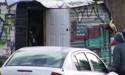 Graffiti-covered van with its back door open, a white car in the foreground, and people in hoodies nearby.
