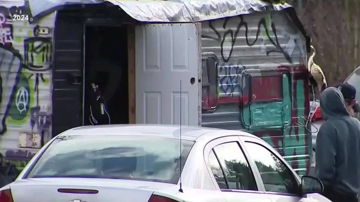 Graffiti-covered van with its back door open, a white car in the foreground, and people in hoodies nearby.
