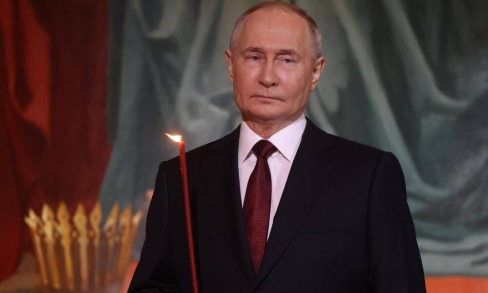 Older man in a dark suit and burgundy tie holds a lit red candle at a formal ceremony.