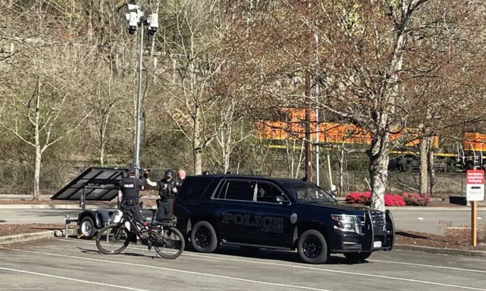 Two police officers stand near a dark SUV marked Police, beside a trailer with a solar panel in a parking lot near train tracks.