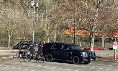 Two police officers stand near a dark SUV marked Police, beside a trailer with a solar panel in a parking lot near train tracks.