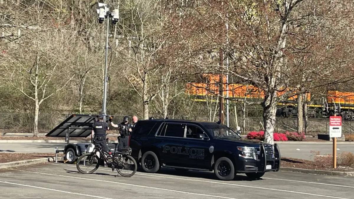 Two police officers stand near a dark SUV marked Police, beside a trailer with a solar panel in a parking lot near train tracks.