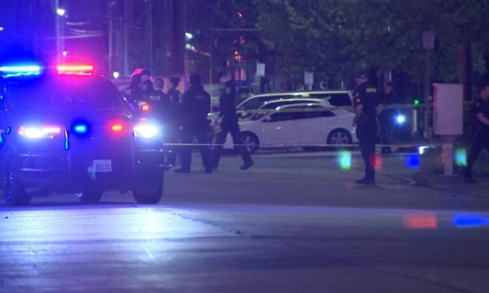 Nighttime police scene with officers, patrol cars, and flashing blue and red lights behind police tape on a street barrier.