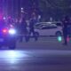 Nighttime police scene with officers, patrol cars, and flashing blue and red lights behind police tape on a street barrier.