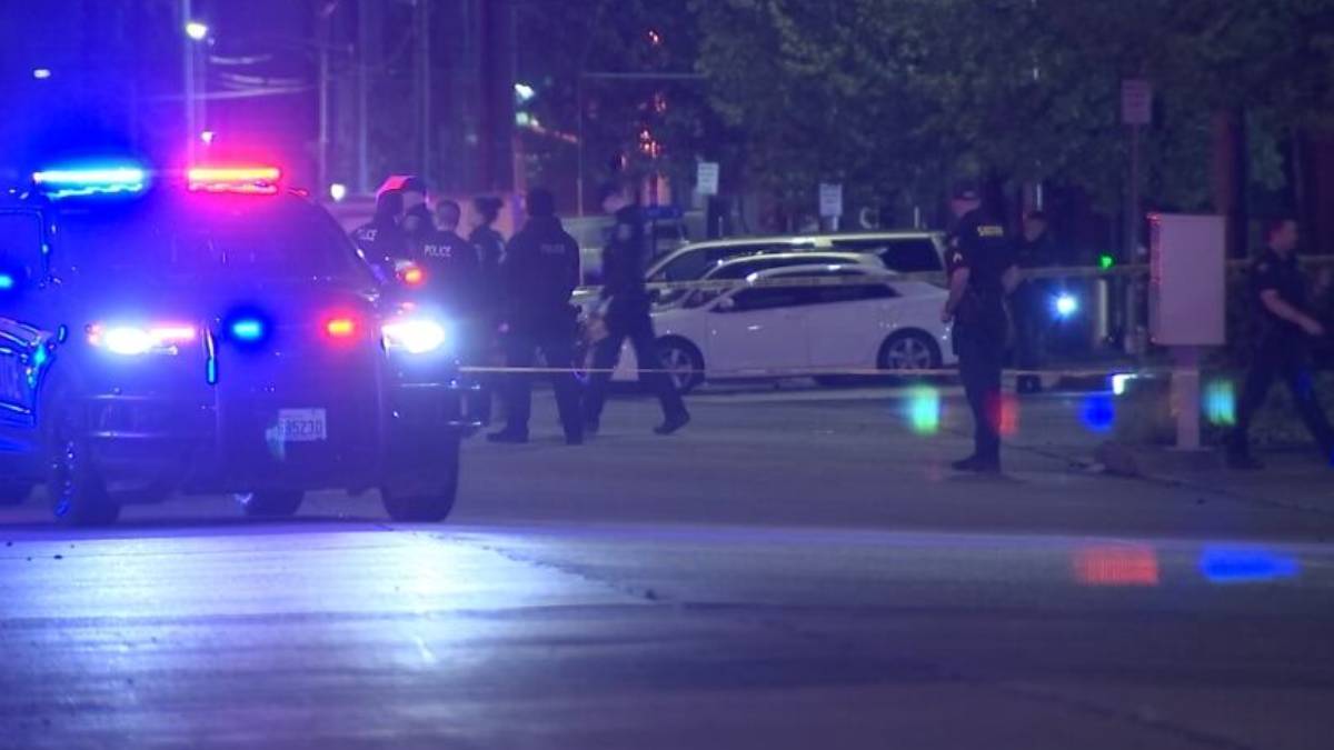 Nighttime police scene with officers, patrol cars, and flashing blue and red lights behind police tape on a street barrier.